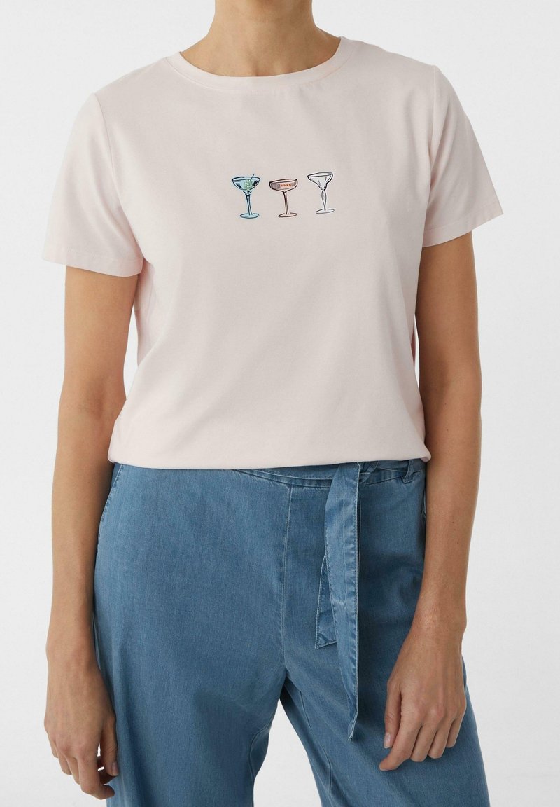 Femme portant un t-shirt rose clair avec trois illustrations colorées de verres à cocktail sur la poitrine et un pantalon bleu avec ceinture nouée.