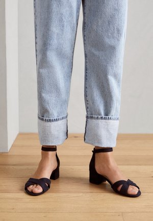 Sandales noires en suède à talons bas avec des brides entrecroisées, associées à un jean en denim bleu clair à revers. Fond en parquet.