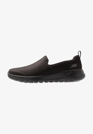 Zapatilla negra sin cordones con parte superior de malla transpirable, suela gruesa acolchada y lengüeta en el talón, mostrada en perfil lateral sobre fondo blanco.