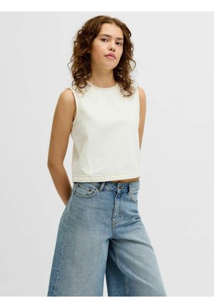 Fiatal nő göndör hajjal, fehér ujjatlan felsőt és bő, világoskék farmert visel egy egyszínű háttér előtt.