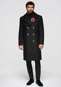 Schwarzer Mantel aus Wollmischung mit doppelreihiger Knopfleiste, goldenen Knöpfen, großen Revers und einer dekorativen roten Herzbrosche, kombiniert mit eng geschnittenen schwarzen Hosen und Stiefeln.