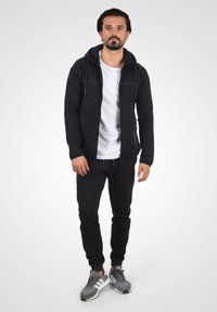 Sort atletisk hættetrøje med lynlåslommer, båret over en hvid t-shirt, parret med sorte joggingbukser og grå sneakers. Har lynlåslukning foran og snore til justering.