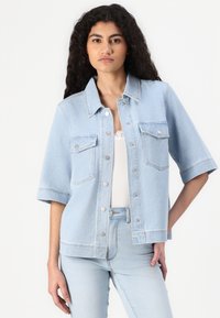 Jaqueta de denim azul claro, mangas curtas, botões de pressão, dois bolsos no peito, design ligeiramente ajustado, com uma textura suave e casual.