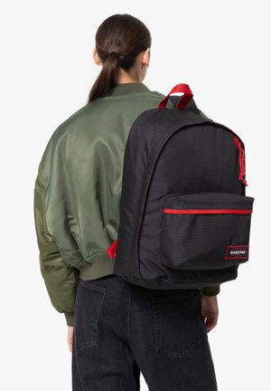 Black fabric backpack with red zipper and front pocket, featuring a textured pattern, worn on the back of a person in an olive green bomber jacket.