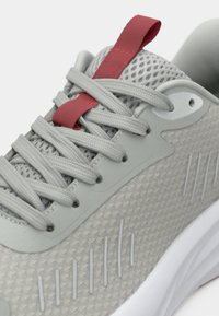 Gaiši pelēks sporta apavu ar acs materiāla augšdaļu, sarkanu akcenta cilpu, teksturētu dizainu, plakanu pelēku šņori un baltu viduspēdu ar smalkām svītrām.