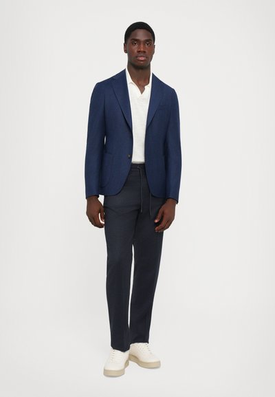 Sacou bleumarin cu două buzunare, asortat cu pantaloni închiși la culoare și teniși crem. Cămașa este de culoare deschisă și are guler. Design și textură simple.
