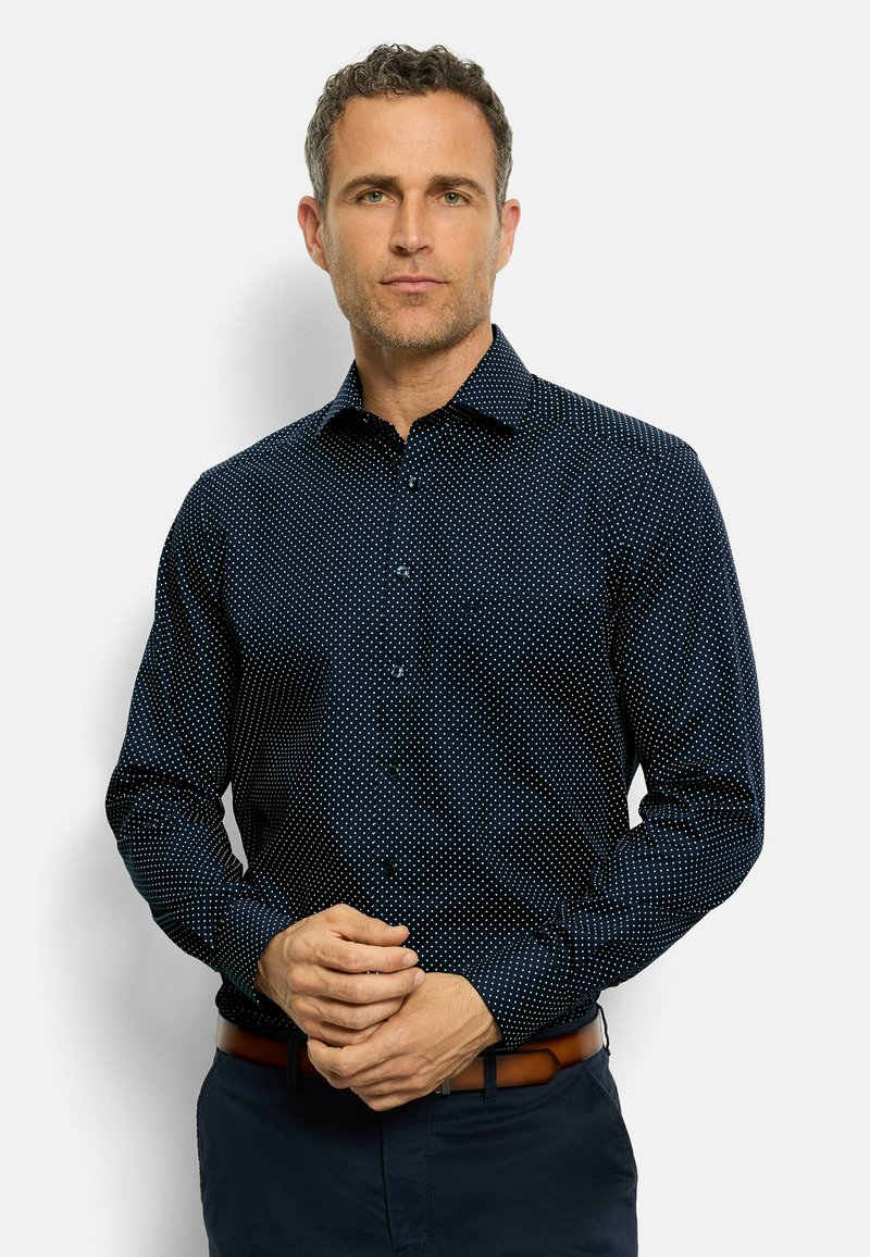 Camicia blu navy con pois bianchi, maniche lunghe, tessuto morbido e colletto classico. Indossata con una cintura marrone e pantaloni scuri.