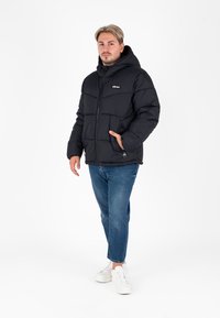 Zwarte gewatteerde jas met een capuchon, ritsluiting, zijzakken en een logo op de borst. G gedragen over blauwe jeans en witte sneakers.