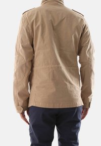 Giacca militare beige con colletto alto, spalline e polsi abbottonati; cuciture delle spalle e toppe sui gomiti visibili.