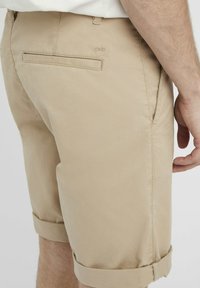 Shorts beiges en tissu à la texture lisse. Présentent des ourlets retroussés, des poches latérales et une poche arrière avec un détail de couture.