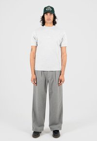 Uomo con lunghi capelli che indossa un cappello verde, una camicia grigia chiara a maniche corte, pantaloni grigi larghi e scarpe nere, in piedi contro uno sfondo neutro.