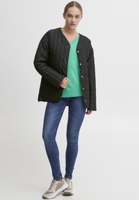 Svart quilted jakke med trykknapper, brukt over en grønn V-hals genser. Kombinert med blå skinny jeans og beige joggesko.