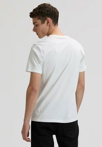 Άσπρο βαμβακερό μπλουζάκι με κοντά μανίκια, στρογγυλή λαιμόκοψη και λεία υφή. Το μοντέλο είναι γυρισμένο πλάτη, συνδυασμένο με μαύρο τζιν.