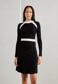 Musta ribbed-mekko pitkillä hihoilla, jossa on valkoisia yksityiskohtia kaula-aukon ja vyötärön ympärillä sekä kultaiset nappitiedot hartioissa.
