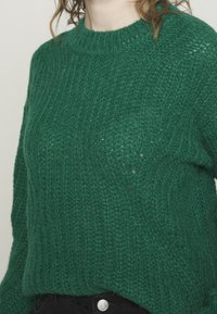 Maglione verde a maglia grossa con scollo a coste indossato sopra pantaloni neri con chiusura a bottone visibile.