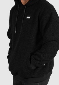 Svart fleecehoodie med en texturerad yta, justerbar dragskohet, och en liten logotypbricka på framsidan. Har framfickor.