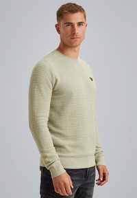 Suéter texturizado verde claro con cuello redondo, puños acanalados y un pequeño logo en el pecho. Combinado con jeans oscuros.