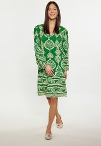 Rochie verde cu model, cu mâneci lungi, având desene florale albe complexe și benzi horizontale. Purtați cu pantofi bej.