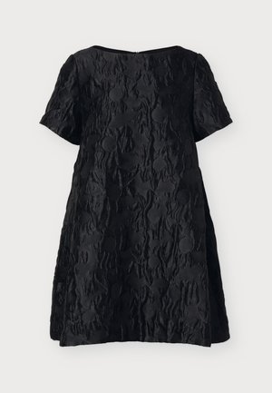 Jeune femme debout portant une courte robe noire à motifs texturés, des chaussettes blanches et des mocassins noirs à pompons sur un fond clair uni.