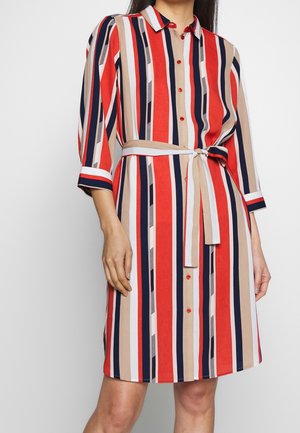 Kobieta w sukience sięgającej do kolan, z długim rękawem, z pionowymi paskami w kolorze czerwonym, beżowym, granatowym i białym, z przodu zapinanej na guziki i z wiązanym paskiem w talii.