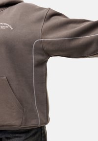 Ruskea huppari valkoisilla saumoilla, jossa on etutasku ja väljä istuvuus. Pehmeä materiaali, rento tyyli ja minimalistiset yksityiskohdat.