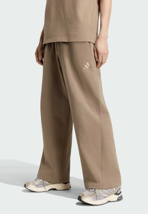 Person iført løstsiddende brune Adidas joggingbukser og hvide, beige og grå sneakers, stående mod en ensfarvet baggrund.