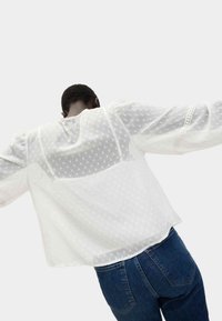 Blusa bianca, con texture e motivo a pois, maniche lunghe trasparenti e taglio leggermente corto, abbinata a jeans blu di denim.