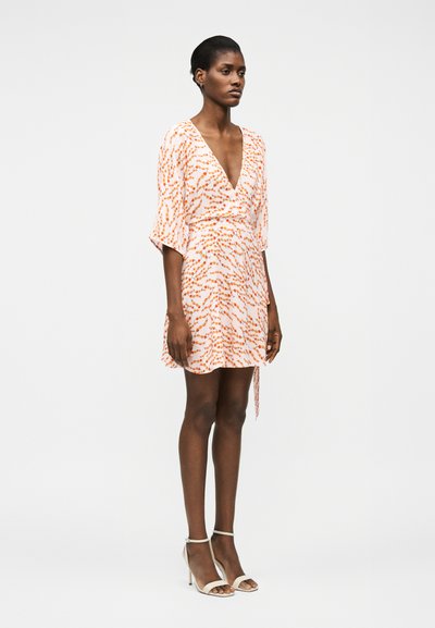 Γυναίκα όρθια που φοράει ένα λευκό και πορτοκαλί φόρεμα τύπου wrap με μανίκια μέχρι τον αγκώνα και λευκά σανδάλια με ψηλό τακούνι.