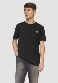 Zwarte katoenen T-shirt met een ronde hals en korte mouwen, voorzien van een klein merklogo op de borst. Gekenmerkt door grijze jeans.