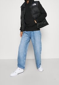 Gilet imbottito nero sopra una felpa nera con cappuccio, abbinato a jeans larghi azzurri e sneakers bianche con dettagli neri.