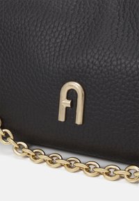 Surface en cuir noir texturé avec un logo métallique doré en forme de « A » abstrait et une bandoulière chaîne dorée attachée.