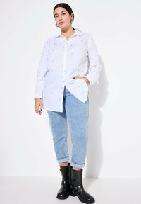 Weißes Langarmhemd mit kleinen silbernen Herzmustern, Knopfleiste und lässiger Passform, kombiniert mit hellblauen Jeans und schwarzen Stiefeletten.