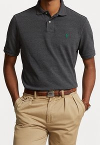 Mörkgrå pikétröja med korta ärmar, dubbelknapplista och grön logotyp. Kombinerad med beige byxor och ett brunt läderbälte.