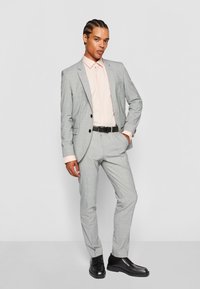 Traje gris claro con un sutil patrón de cuadros, combinado con una camisa rosa pálido, cinturón negro y zapatos negros, de pie contra un fondo blanco.