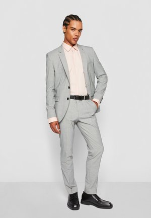 Lichtgrijs pak met subtiel ruitpatroon, gecombineerd met een lichtroze shirt, zwarte riem en zwarte schoenen, staande tegen een witte achtergrond.