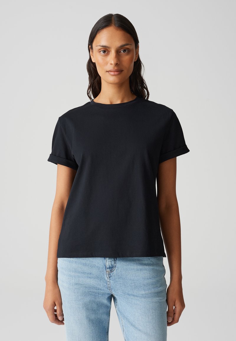 T-shirt di cotone nero con maniche corte arrotolate, collo a giro, vestibilità rilassata, indossato con jeans in denim blu chiaro, texture liscia.