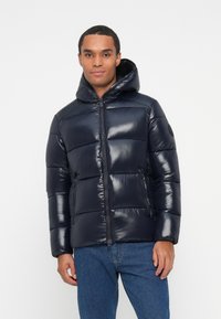 Man wearing a shiny dark blue puffer jacket with hood and front zipper, paired with blue jeans, standing against a plain white background.