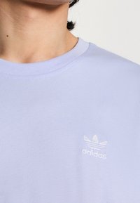 Světle fialové tričko, bavlněná tkanina, kulatý výstřih, s malým bílým vyšitým logem Adidas. Hladká textura, minimalistický design.