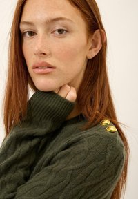 Zielony sweter w splot kablowy z prążkowanymi mankietami i trzema złotymi guzikami na ramieniu. Miękka tekstura, dopasowany krój.
