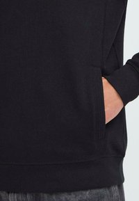 Mano nella tasca di una felpa nera con cuciture visibili, manica a polsino e parte di jeans grigi consumati.