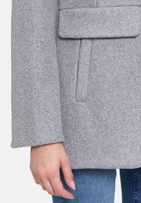 Grau melierter Wollmantel mit einem schlichten Design, großen Front-Pattentaschen und weicher Textur. Das Modell trägt hellblaue Jeans.