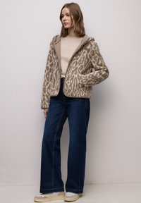 Beige flauschige Jacke mit Leopardenmuster, Reißverschluss und Kapuze, kombiniert mit einem hellen Oberteil, ausgestellten dunklen Jeans und cremefarbenen Sneakers.