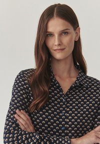 Blusa blu navy con motivo floreale bianco e bottoni blu; presenta un collo a camicia e maniche lunghe con polsini abbottonati.