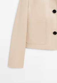 Beige kunstlæderjakke med lige skæring, lange ærmer, sorte knapper og en patch-lomme på venstre side. Glat tekstur.