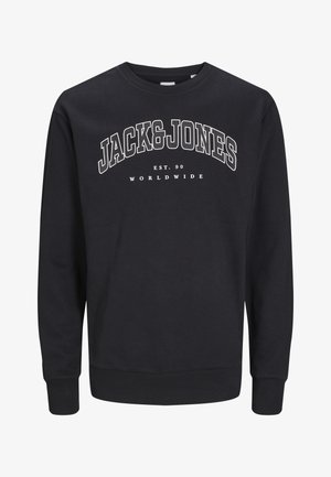 Blond chłopiec stoi z rękami wzdłuż ciała, ubrany w czarną bluzę Jack & Jones, czarne spodnie typu cargo i brązowe trampki na jednolitym tle.