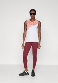 Femme portant un haut de sport sans manches blanc et orange, un legging bordeaux avec un logo, des baskets noires et des lunettes de soleil de sport sombres, se tenant devant un fond neutre.