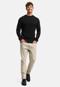 Schwarzer Kabelstrickpullover, beige Hose und schwarze Schuhe. Glatte Stofftextur des Pullovers; taillierte Passform mit gerippten Bündchen.