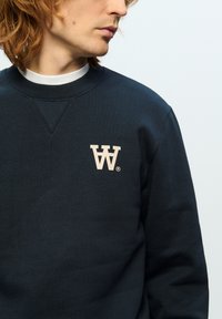 Dark navy sweatshirt made of cotton blend, featuring a light beige stylized "W" logo on the left chest and a crew neckline.