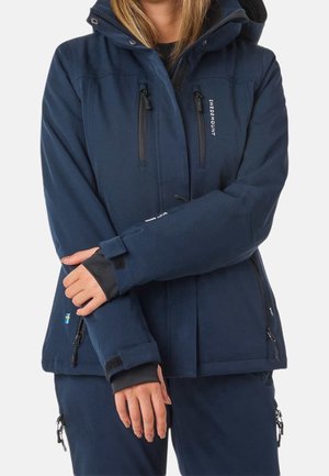 Vrouw die een marineblauwe waterdichte jas draagt met ritssluitingen op de borstzakken en klittenband aan de polsen, houdt één arm over haar lichaam gekruist.
