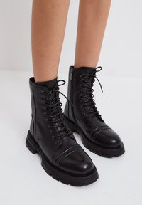 Sorte læderkampstøvler med runde tæer, ribbede detaljer og snørebånd foran. De har en sidelukning med lynlås og en tyk, robust sål.